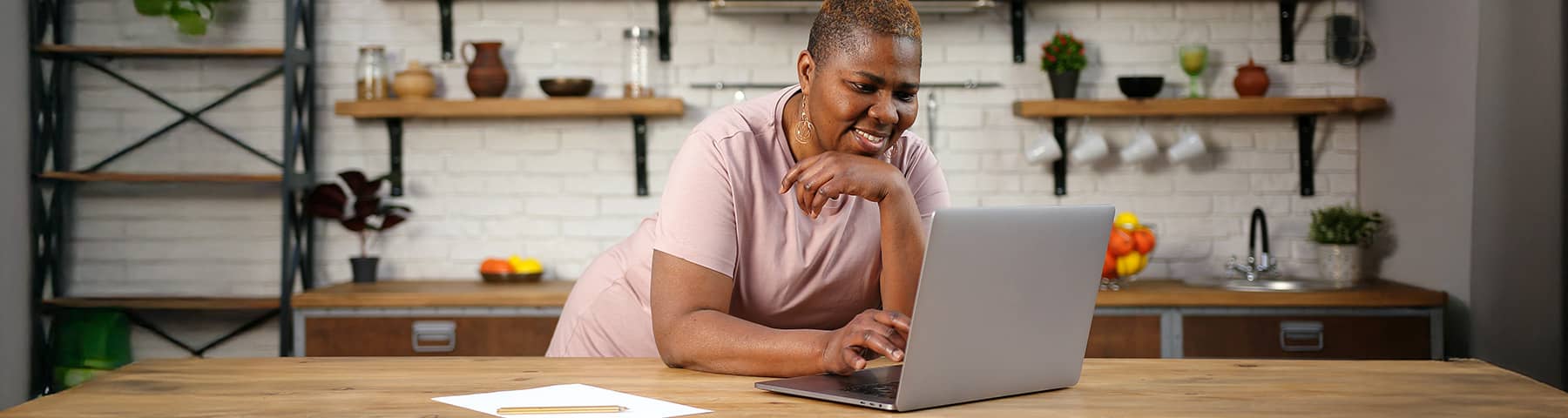 Smiling woman with a short haircut leaning on a kitchen counter, watching a seminar on her laptop.