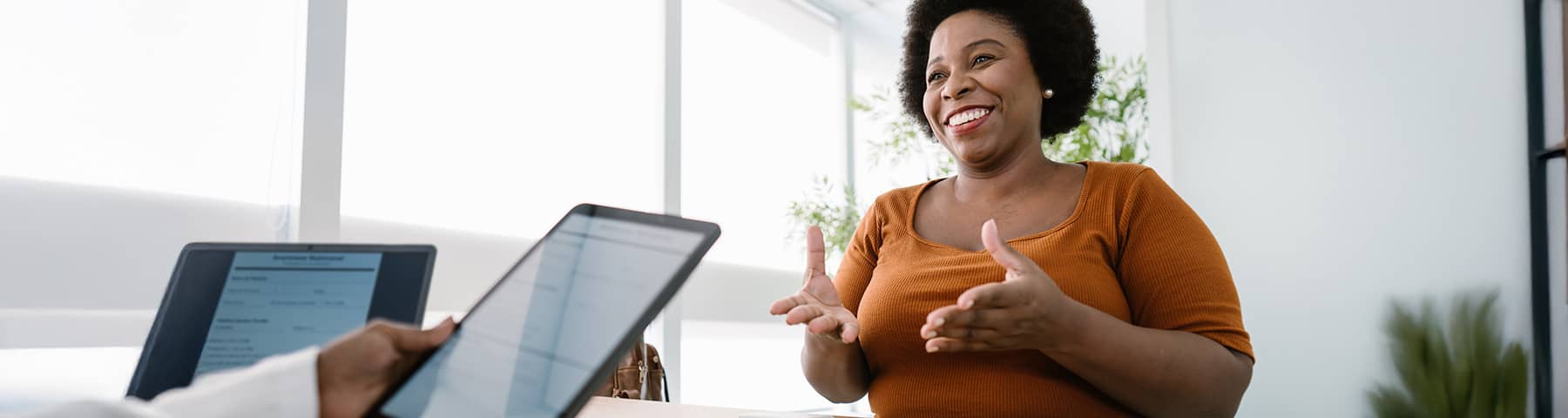 Smiling woman with a larger build wearing an orange top, gesturing while speaking with a healthcare professional holding a tablet.