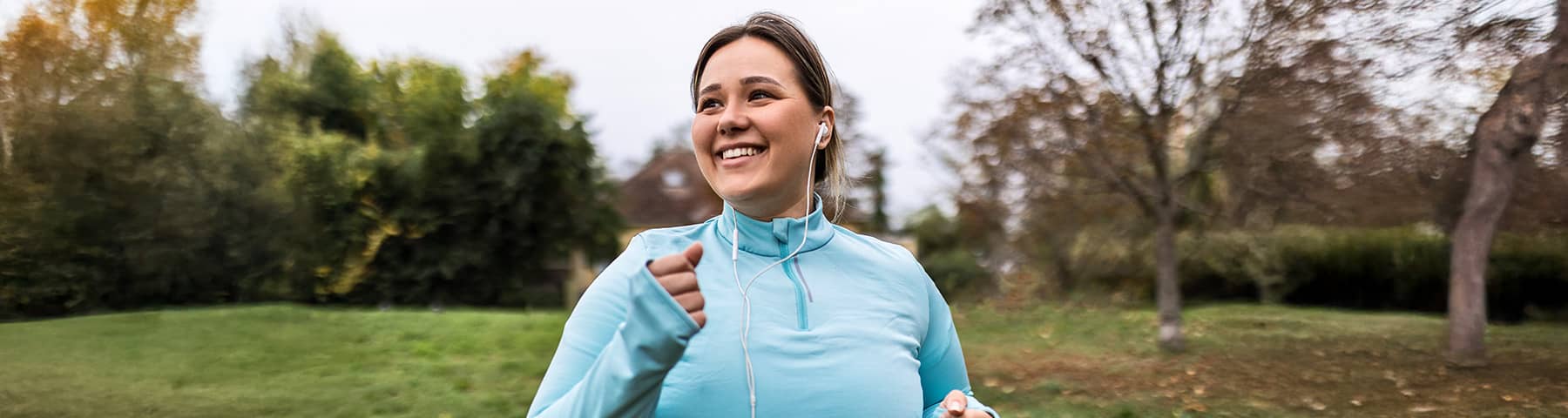 Smiling woman with a medium build jogging outdoors in a light blue athletic top and wearing earphones.