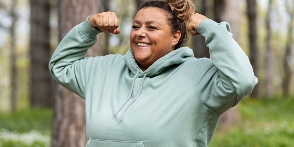 Smiling woman flexing her arms in a green hoodie outdoors.