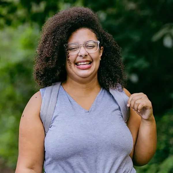 A smiling woman with curly hair and glasses stands outdoors wearing a lavender shirt and a backpack strap.